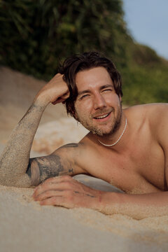 Shirtless tattooed man lying on sand propped on arms smiling at camera