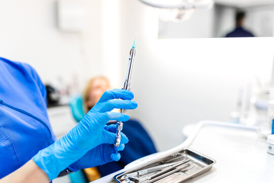 Close up of dentist hand in blue gloves holding anesthesia syringe in dental clinic. Professional medical equipment for pain relief during surgery. Dentist office background with patient blurred.