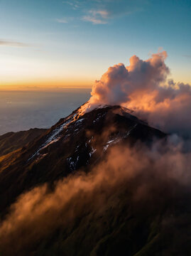 Aerial View of Active Volcano Crater With Rising Smoke and Steam Illuminated by Orange Sunset Light Above Cloud Layer