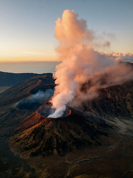 Aerial View of Active Volcano Crater With Rising Smoke and Steam Illuminated by Orange Sunset Light Above Cloud Layer