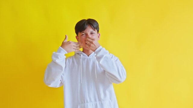 Teenage boy showing disgusted expression due to foul stink gesturing with hand against yellow background