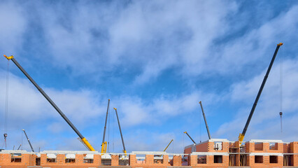 Panoramic view of a brick house construction site with several yellow telescopic cranes working under a blue sky with soft clouds. Industrial building process of a multi-story apartment block. © Анатолий Еремин