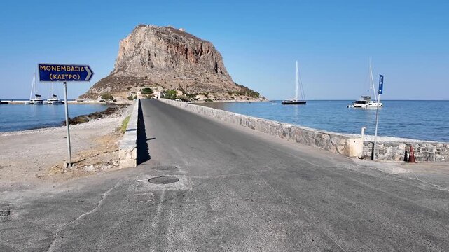 Peloponnese,Monemvasia,Still Frame of the bridge road to the old citadel surrounded by sea on both sides.Big rock formation of the citadel outstanding on a sunny day.Boats on anchor.