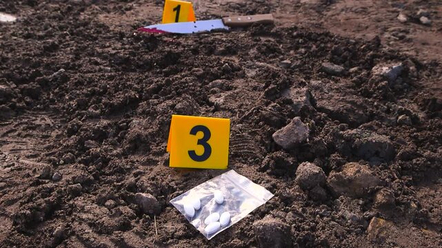 Close up footage of yellow numbered Crime scene markers next to murder evidence like a bloody knife, white colored pills drugs and a footprint on a muddy road outside. Knife has bloodspatter on it.