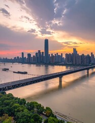 Obraz premium A stunning aerial view captures a modern cityscape illuminated by a vibrant sunset, with a bridge gracefully spanning a wide river.