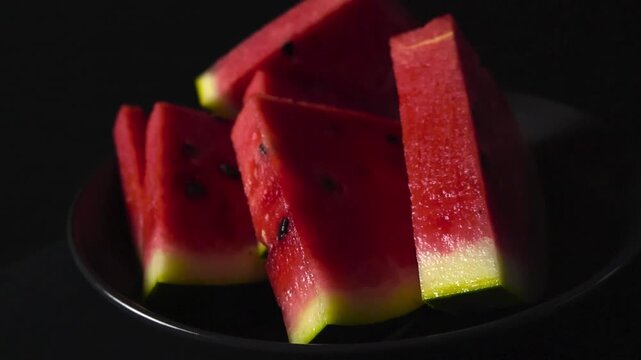 Close up video orbiting around fresh colorful and delicious watermelon triangle shaped slices that are in a bowl in front of bokeh blurry black studio background. Seeds visible in the fruit.