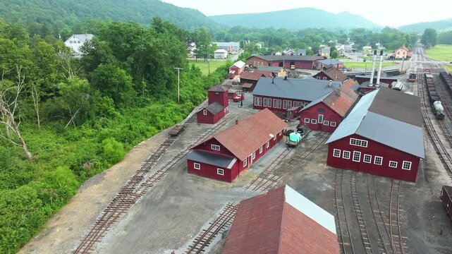 Visit a charming train yard surrounded by mountains. Discover red wooden buildings, rail tracks, and lush greenery. Experience the quiet beauty of this historical site on a sunny day.