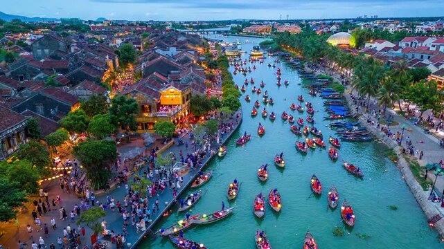 Aerial hyperlapse of traditional wooden boats with lanterns on the Thu Bon River and crowds walking through the historic district at dusk in Hoi An, Vietnam.
