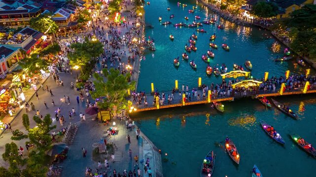 Aerial hyperlapse of adult men and women on small boats with lanterns near a crowded bridge over the Thu Bon River in Hoi An, Vietnam.