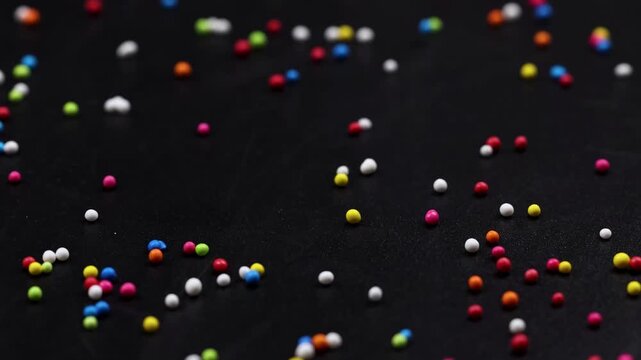 Colorful candy sprinkles falling on black background slow motion, vibrant round sugar confectionery for dessert decoration.
