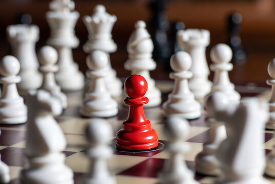 A single red pawn stands out on a chessboard with numerous white chess pieces. It represents a lone individual or outlier within a group.
