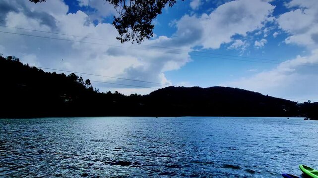 A serene lakescape in Nainital, Uttarakhand, India, showing gently rippling water under a vast blue sky with clouds, where a lone bird glides above silhouetted hills and faint boats drift in distance.