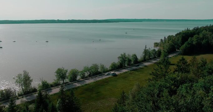 Aerial view of small van driving on road along shoreline of large lake with forest and sailboat in distance
