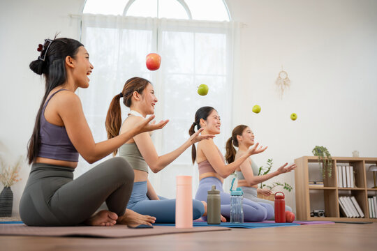 Group of happy Asian women juggling apples and laughing after yoga class at home, Cheerful female friends in sportswear playing with green and red apples during workout break