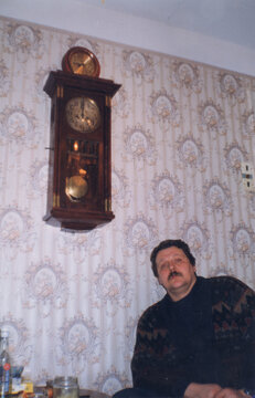 Middle aged man seated in armchair in domestic interior with large vintage pendulum wall clock above him, analog color film mid 1990s