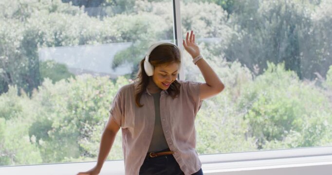 African American woman raising arm, swaying, singing at glass window with music in white headphones