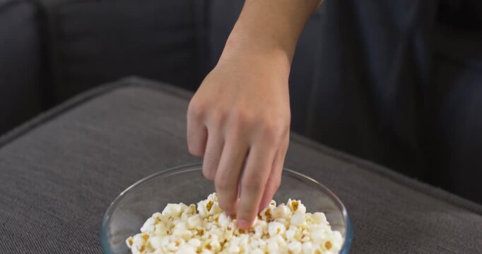 Asian man sitting on dark-gray couch, using smartphone and snacking on popcorn from clear bowl
