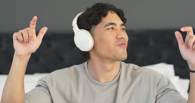 Asian man on bed listening to music with white headset, lifting fingers, forming X, mouthing lyrics