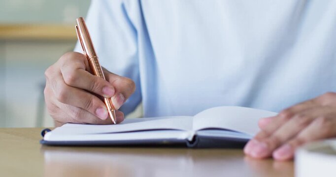 Man holding bronze pen hovering above left page, writing notes and steadying notebook for study
