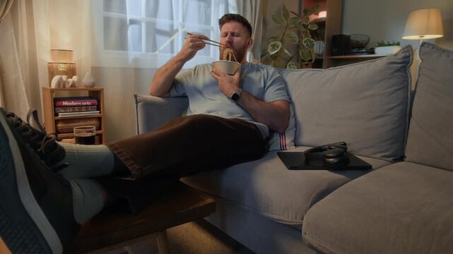 POV full slowmo shot of young Caucasian man relaxing on couch near laptop and headphones in modern living room and eating noodles with chopsticks from takeaway bowl in evening at home