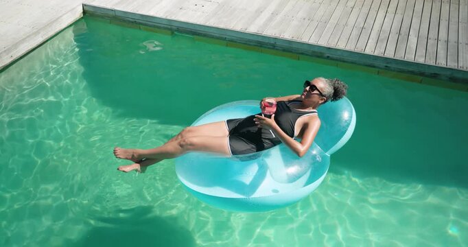 African American woman sunbathing on float in pool sipping pink drink balancing after raft drift