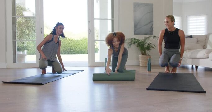 Three women kneeling in home studio, center unrolling green mat, peers aligning for floor routine