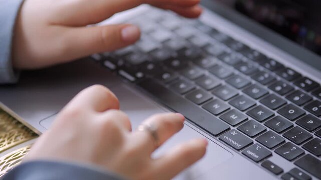 Close-up of hands typing on laptop keyboard. Fingers press keys with precision. One hand wears ring. Background blurred. Laptop silver with black keys