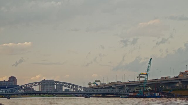 A static shot of the Sumida River in Tokyo, featuring a blue arched bridge and a multi-level expressway under a late afternoon sky.