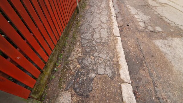 Cracked sidewalk asphalt along a curb beside a red wooden fence, showing gritty freeze-thaw wear after winter&mdash;urban street edge damage and maintenance concept.