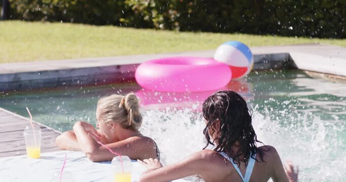 Diverse female friends in swimwear playing at pool as redhead jumping in, splashing cups by phone