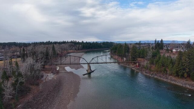 Aerial drone footage of a distinctive steel arch bridge spanning a wide river bordered by pebbled shores, evergreen forests and rural banks near Whitefish, Montana under overcast skies.