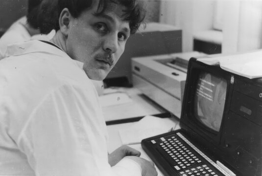 Young man in white lab coat sitting at early computer terminal with keyboard and CRT monitor in laboratory, scanned black and white print circa 1980s