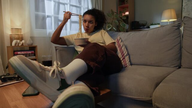 POV full slowmo shot of young African American woman eating noodles with chopsticks while relaxing on sofa spending cozy evening in modern living room