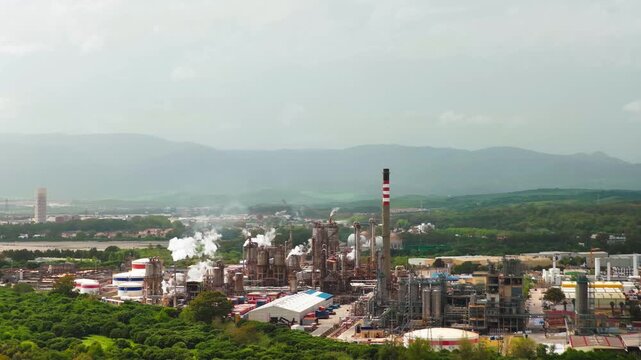 Aerial view of a sprawling industrial complex, plumes of smoke rising against a backdrop of green hills, San Roque, Andalusia, Spain.