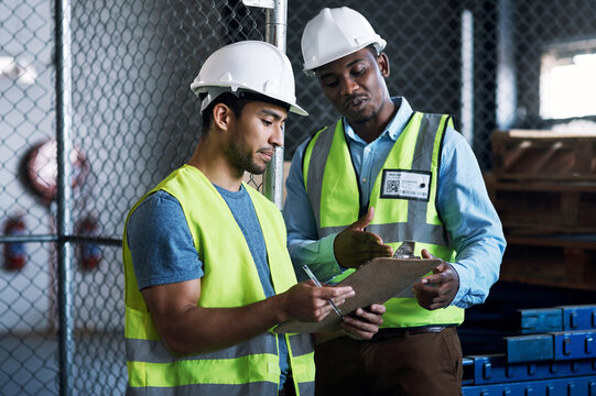 Men, discussion or building contractor with clipboard for checklist or maintenance in warehouse. Male people, employees or conversation with hard hat or plan for construction idea, safety or security