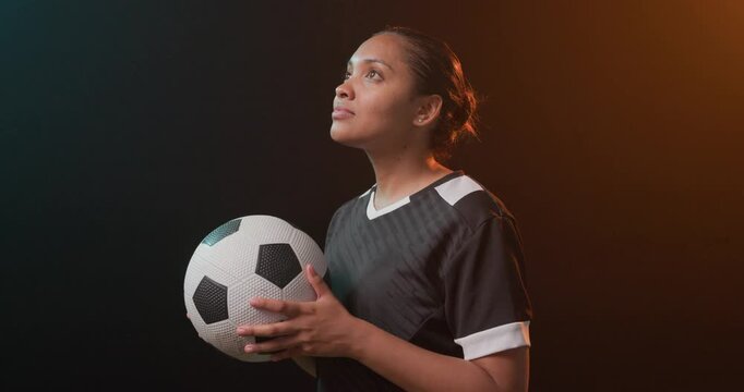 Woman holding soccer ball black jersey studio teal-orange rim, camera closing in, looking up