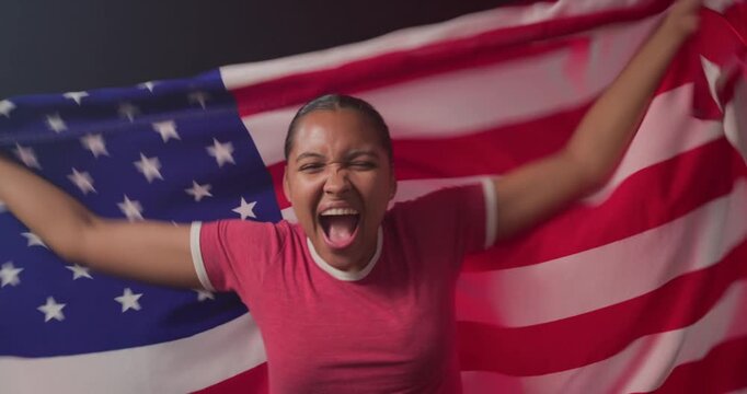 Adult African woman wearing red T-shirt, holding US flag taut and waving while cheering in studio
