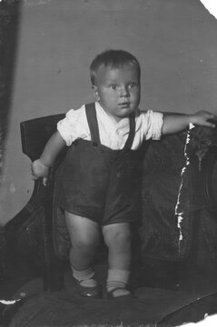 Baby boy in white shirt and dark suspender shorts standing on upholstered armchair, scanned archival black and white print circa 1940s with torn border