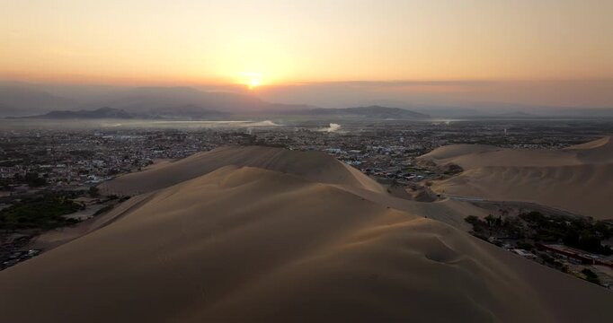 Vast sand dunes surrounding the desert oasis of Huacachina at golden hour sunrise in Ica, Peru. Aerial forward, copy space