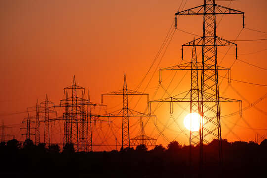 Silhouette of several high-voltage power lines and electricity pylons against a glowing orange and red sunset sky in Germany.