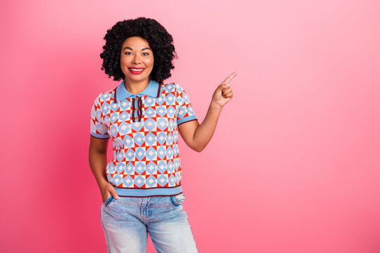 Happy young woman curly hair wear geometric patterned shirt and jeans points right pink background smiling stylish fashion lifestyle advertisement