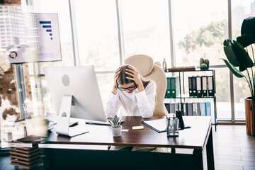 Stressed businesswoman at desk holding her head in a modern office with computer and paperwork,...