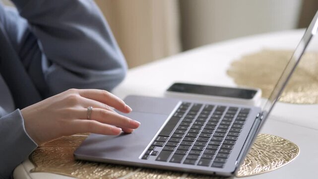 Person works on laptop at round table. Hand moves across touchpad. Phone rests beside laptop. Table has gold textured placemats. Background softly blurred. Light comes from window