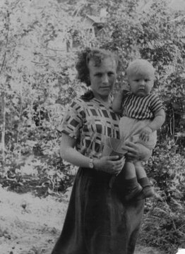 Young woman in patterned dress holding infant in striped shirt in arms outdoors in summer garden with trees, scanned black and white print circa 1950s