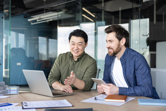 Two diverse male colleagues are smiling while discussing a project, looking at a laptop, and collaborating with a tablet in a modern office environment