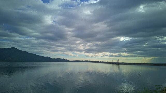 Time lapse stunning landscape view of Khun Dan Prakan Chon Dam showcasing calm blue waters reflecting dramatic white clouds and lush green mountains, creating a peaceful natural panorama.