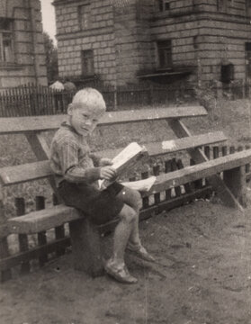 Young boy sitting on wooden bench reading book in yard near brick building, scanned black and white print circa 1950s