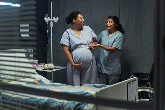 Medium shot of pregnant woman receiving support from nurse in hospital room, holding belly and smiling. Image fitting maternity care, prenatal health, patient support marketing