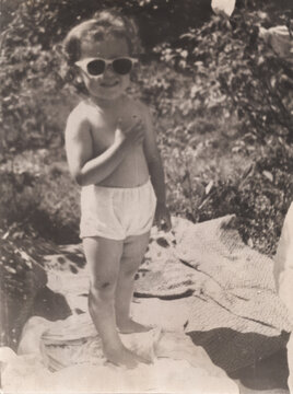 Young girl wearing oversized sunglasses standing outdoors in white bloomers smiling, garden background, scanned black and white print circa 1950s