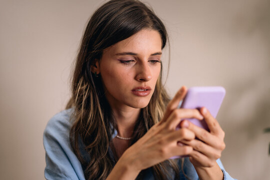 Focused young woman reading smartphone message with serious expression at home. A focused woman in blue blouse uses a lavender smartphone, conveying modern communication and technology.